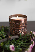 Candle in a copper holder with decorative patterns, surrounded by greenery and flowers.